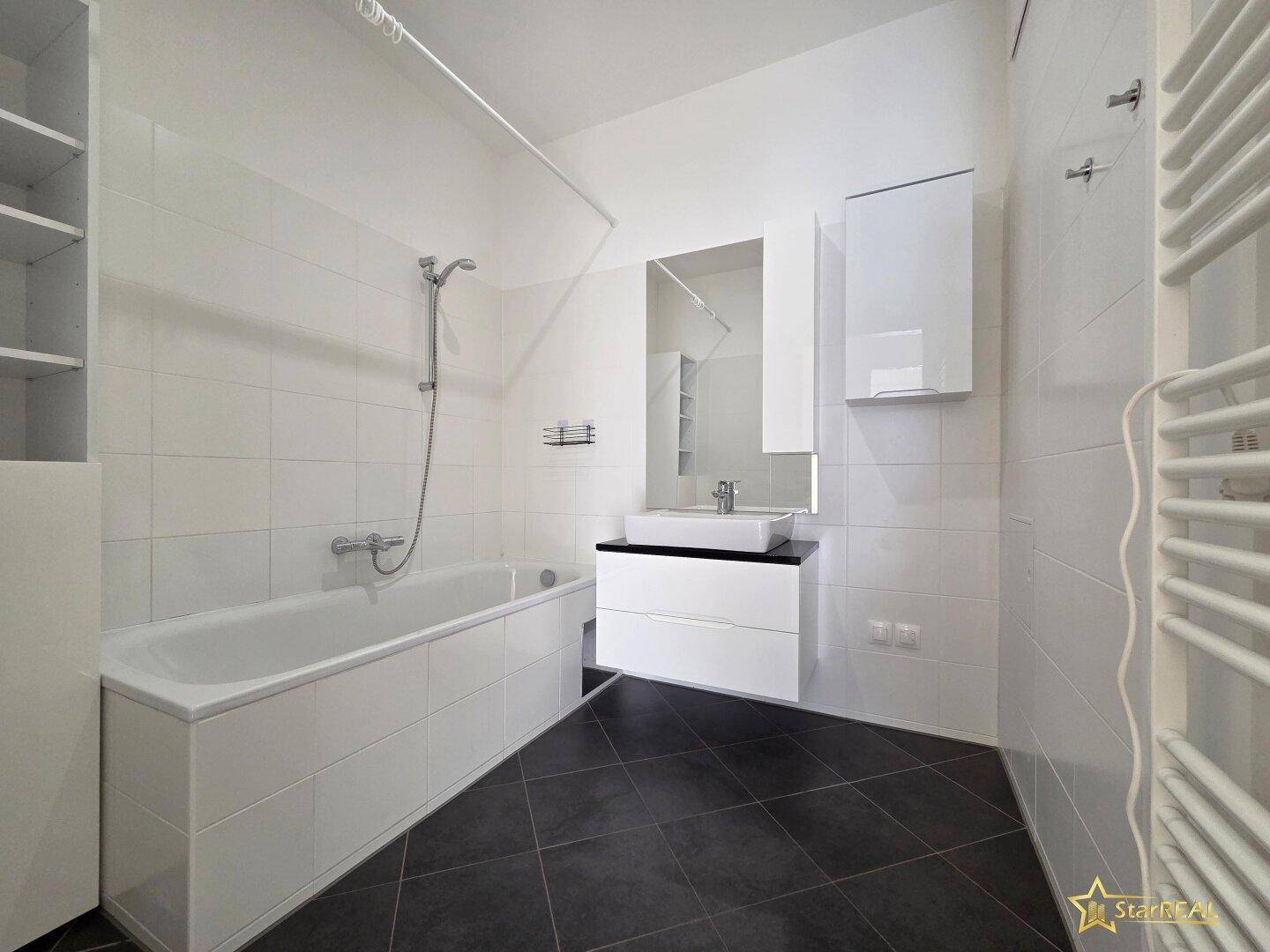 Helles Badezimmer mit Badewanne, modernem Waschtisch und Spiegel, sowie dunklem Fliesenboden.