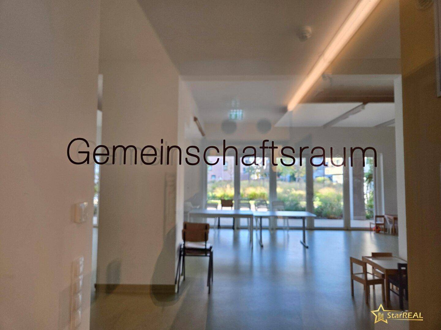 Gemeinschaftsraum mit Tischen und Stühlen, durch eine Glasscheibe sichtbar.