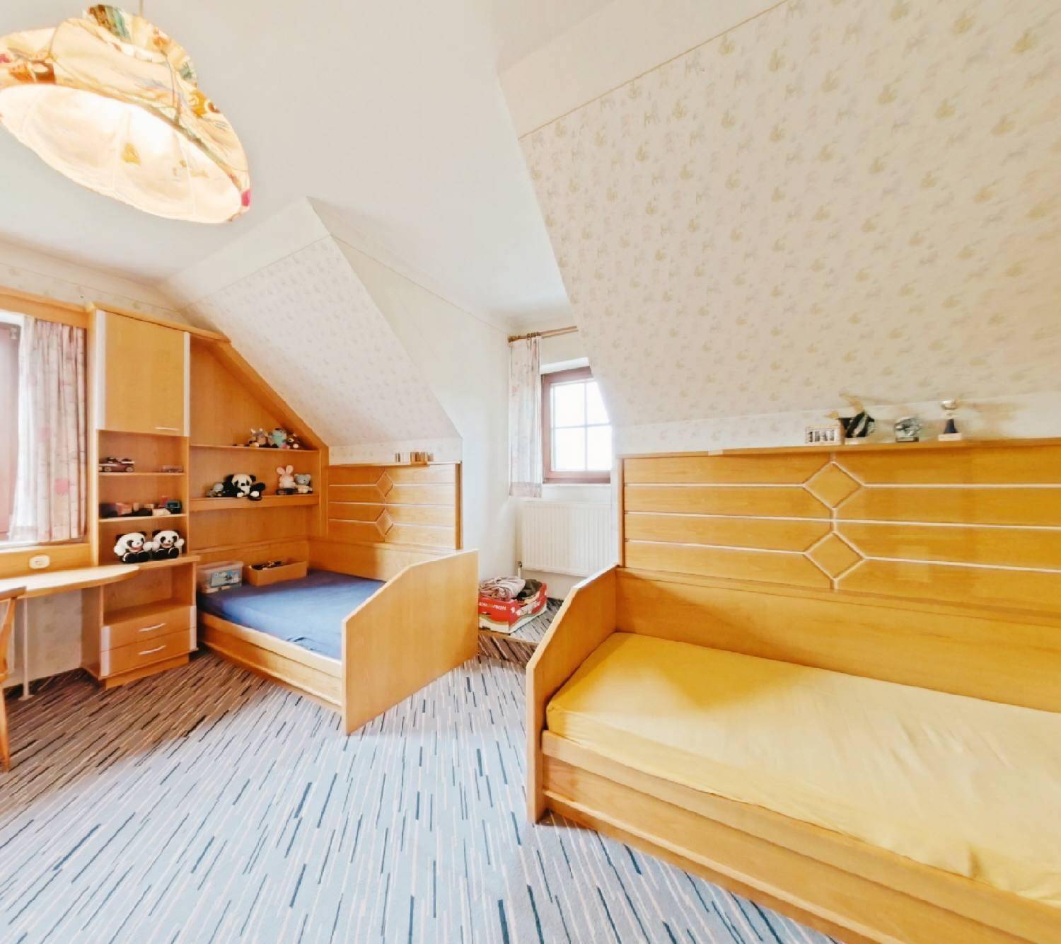 Kinderzimmer mit zwei Holzbetten, Schreibtisch und gemustertem Teppichboden unter der Dachschräge.