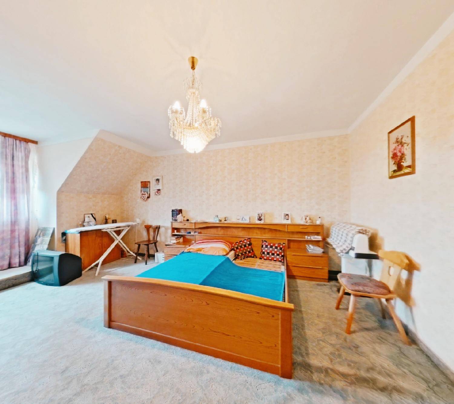 Geräumiges Schlafzimmer mit Holzbett, Teppichboden und einem eleganten Kronleuchter unter der Dachschräge.