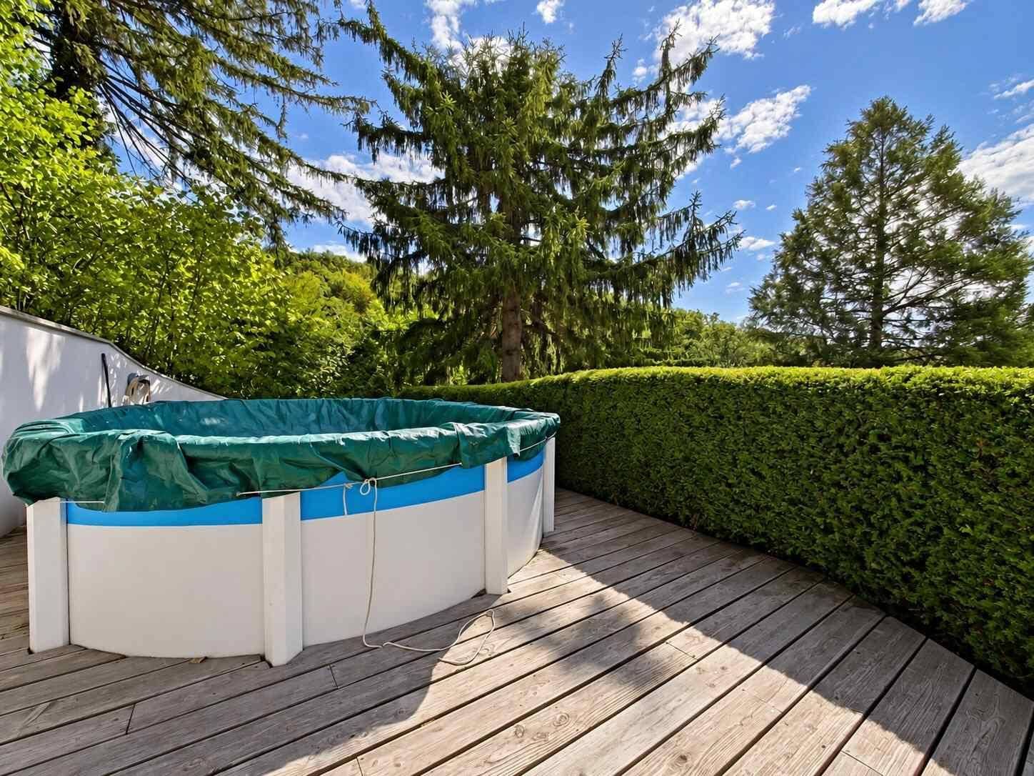 Gartenbereich mit Aufstellpool, Holzterrasse und hohen Bäumen unter blauem Himmel.