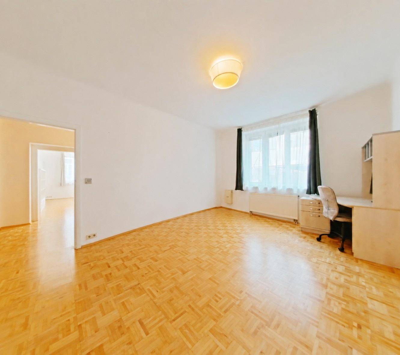 Helles Zimmer mit Parkettboden, Fenster und einem kleinen Arbeitsbereich.