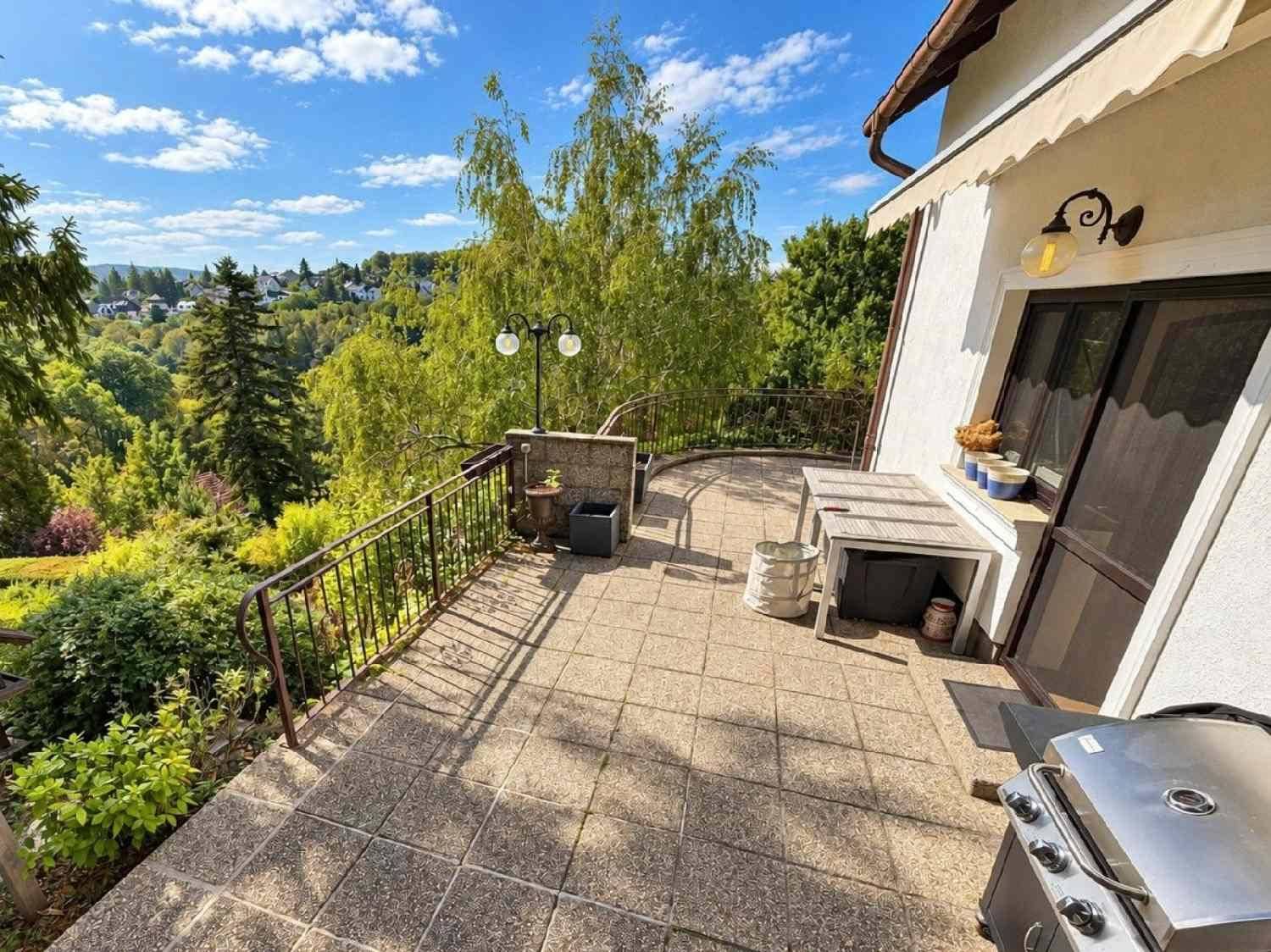 Große gepflasterte Terrasse mit Geländer, Grill und weitem Blick über die bewaldete Landschaft.
