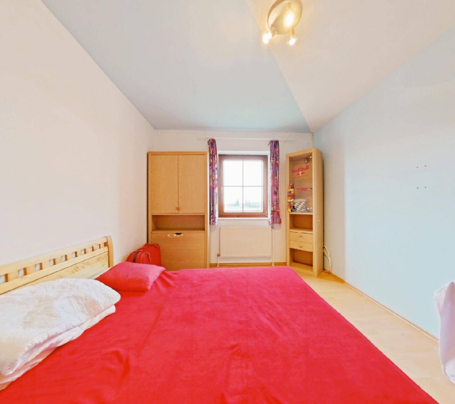 Schlafzimmer mit rotem Bett, Holzfußboden, Kleiderschrank und einem Fenster, das Tageslicht hereinlässt.