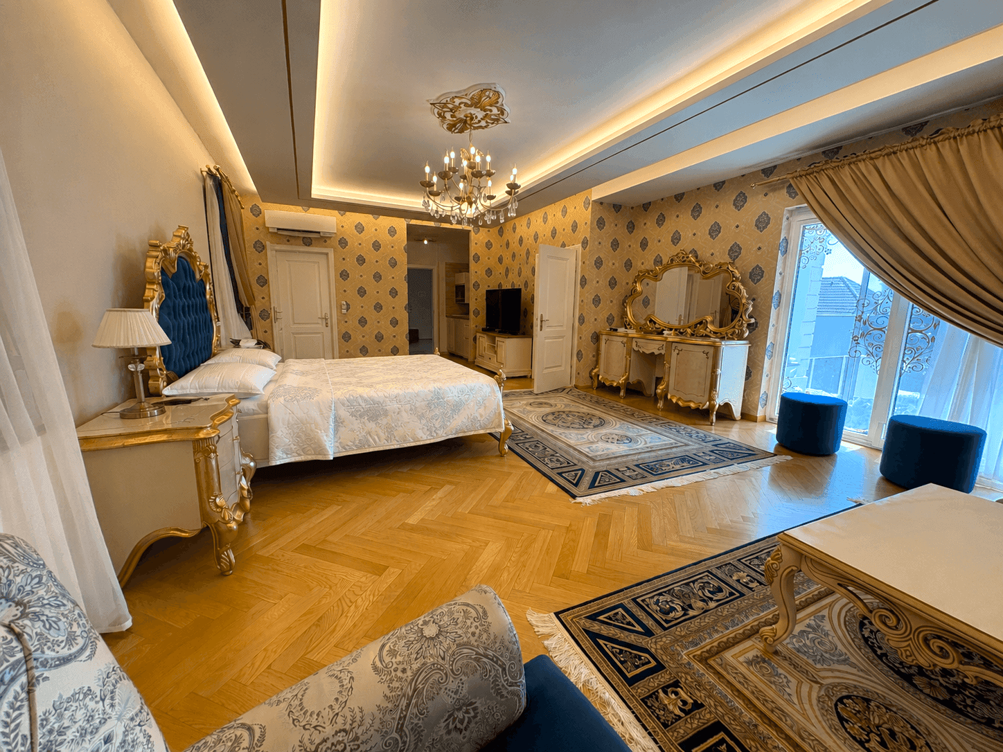 Luxuriöses Schlafzimmer mit opulentem Bett, eleganten Möbeln, gemusterter Tapete und Zugang zu einem Balkon.