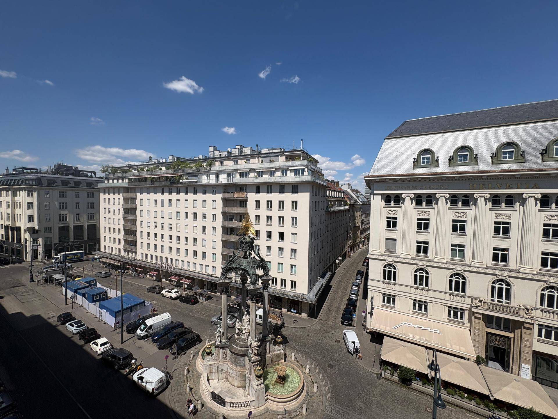 Sonniger Stadtblick auf einen Platz mit einem Denkmal und umliegenden historischen Gebäuden.