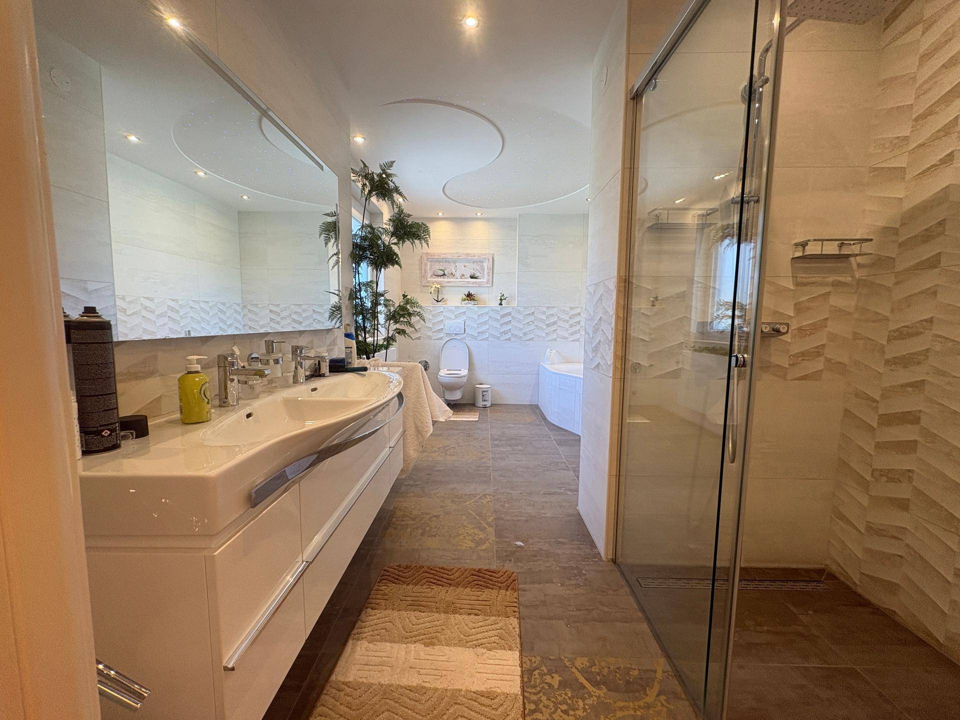 Modernes Badezimmer mit Doppelwaschbecken, großer Spiegelfläche, begehbarer Dusche und Badewanne im Hintergrund.
