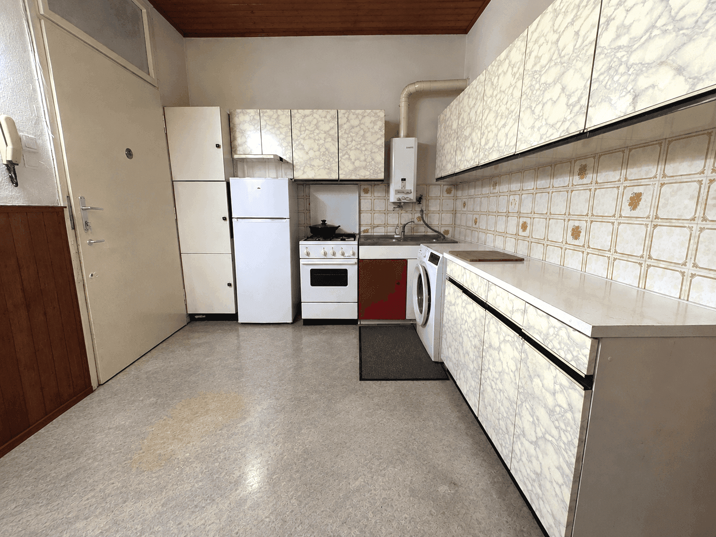 Funktionale Küche mit älteren Schränken im Marmor-Look, Gasherd, Kühlschrank und Waschmaschine.