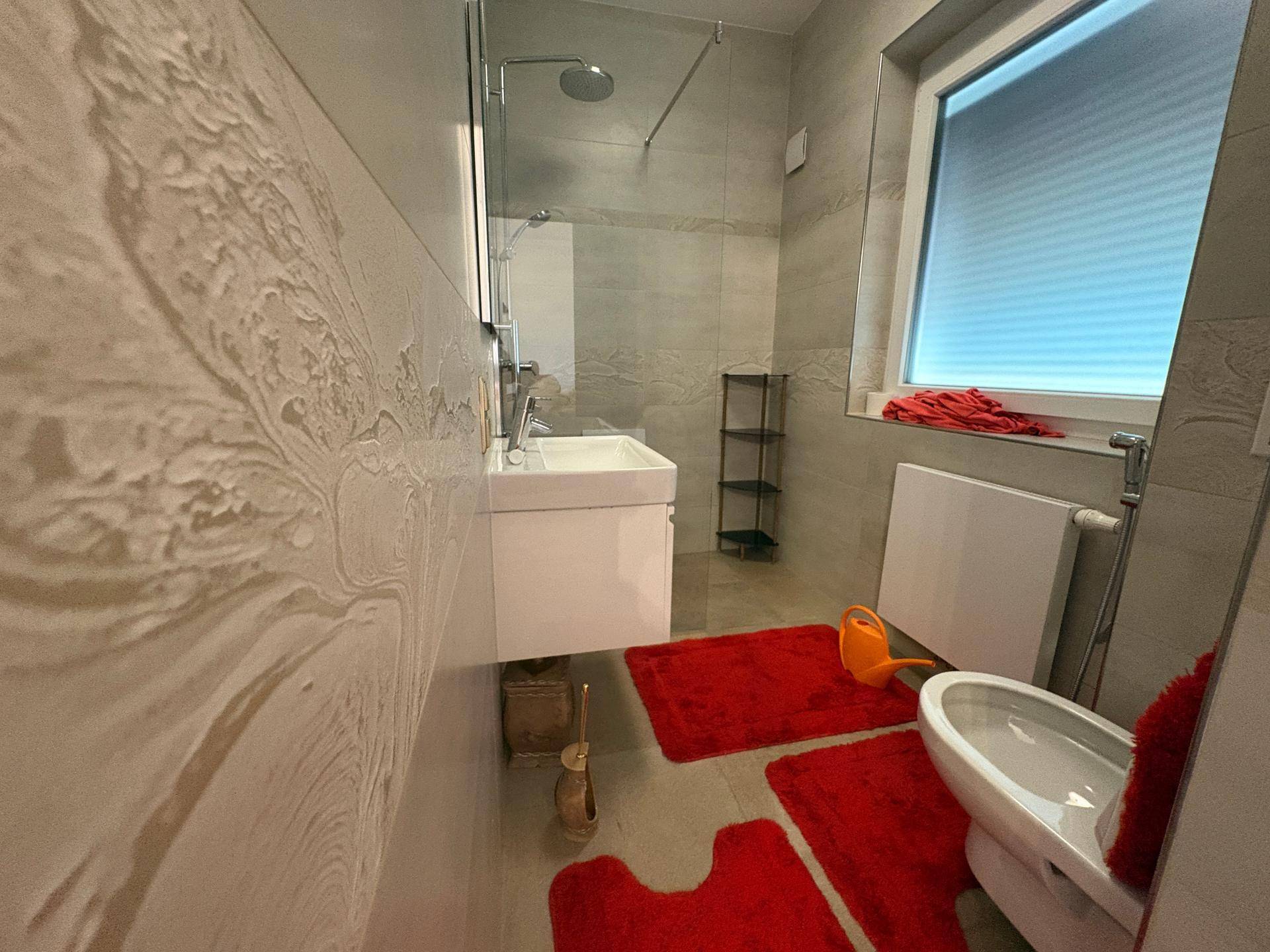 Modernes Badezimmer mit Dusche, wandhängendem Waschbecken und WC, durch ein Fenster hell beleuchtet.