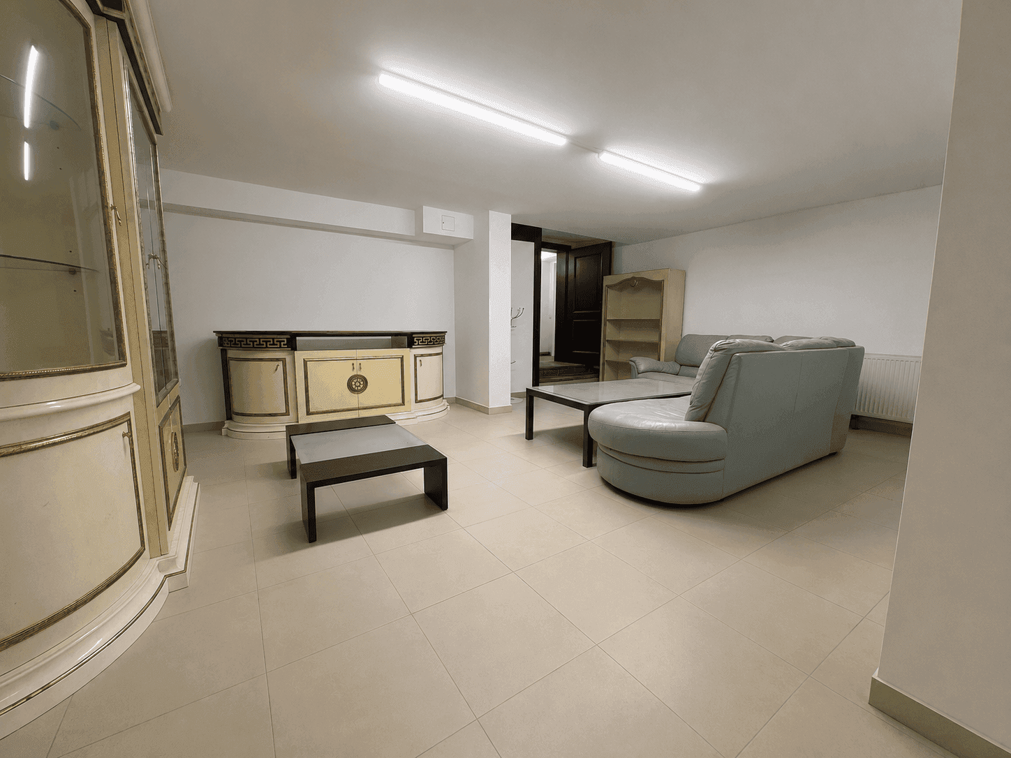Geräumiges Wohnzimmer im Untergeschoss mit Fliesenboden, Sofa und traditionellen Möbeln.