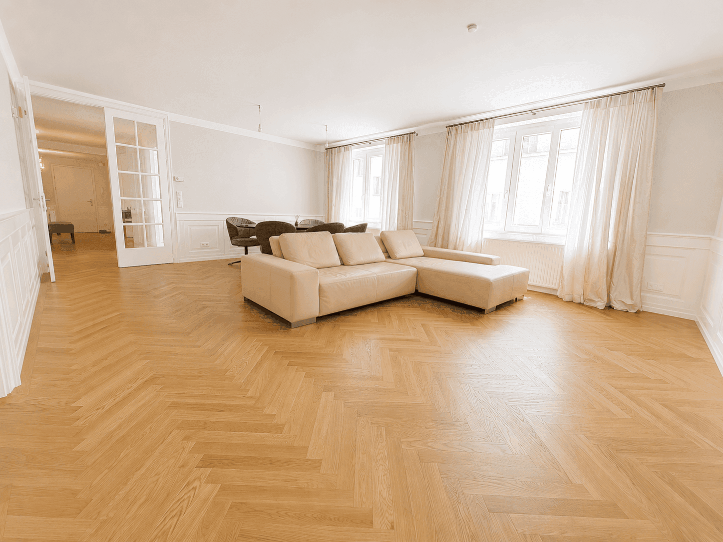 Geräumiger Wohnbereich mit beigem Ecksofa, Fischgrätparkett und eleganten französischen Türen.