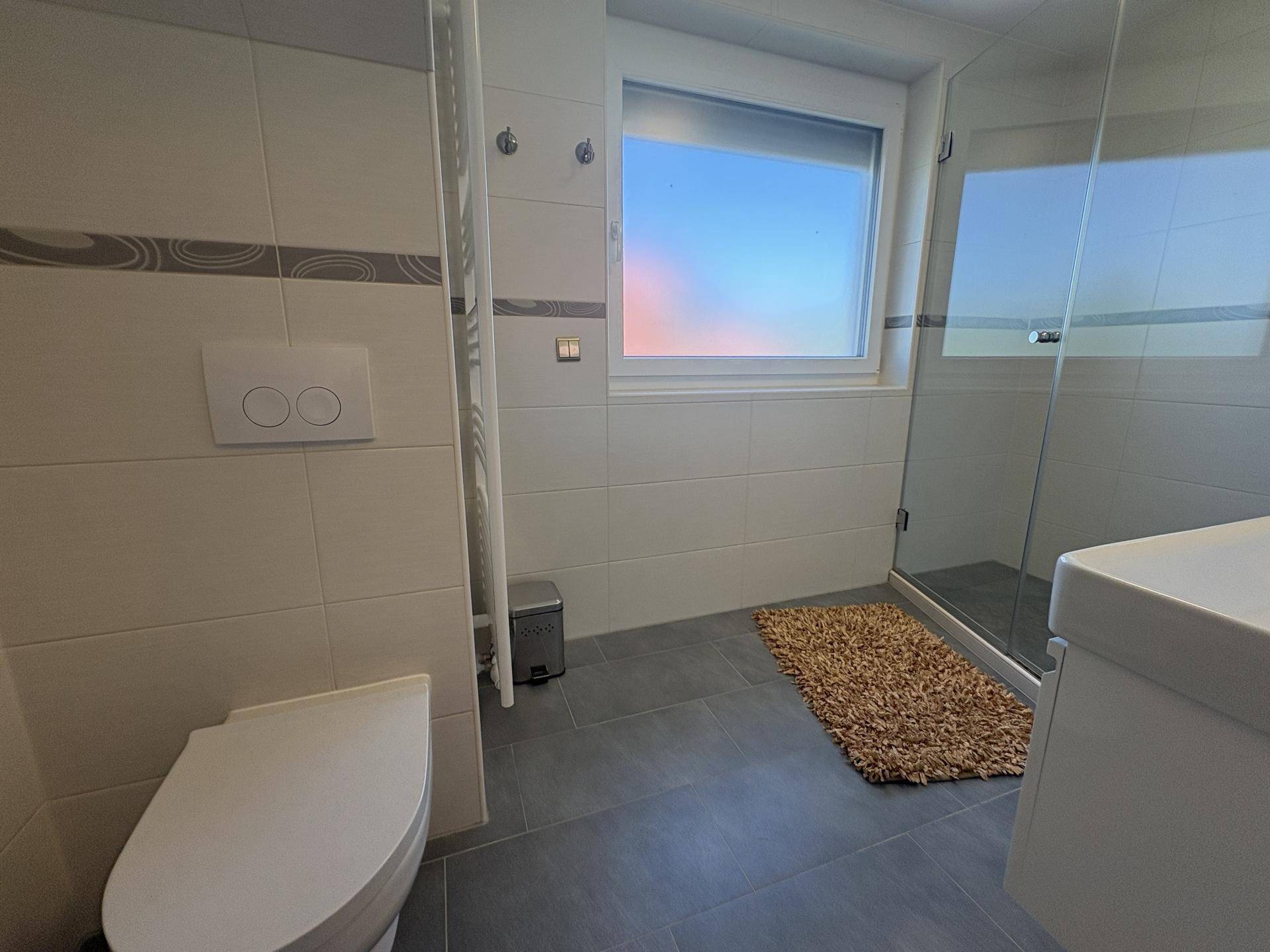Modernes Badezimmer mit WC, Waschtisch, Dusche und Fenster für natürliche Belüftung und Licht.
