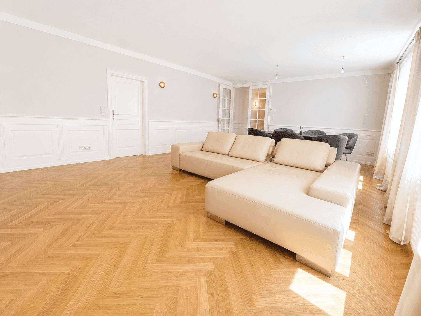 Eleganter Wohnbereich mit beigem Ecksofa, Fischgrätparkett und Blick auf den Essbereich.