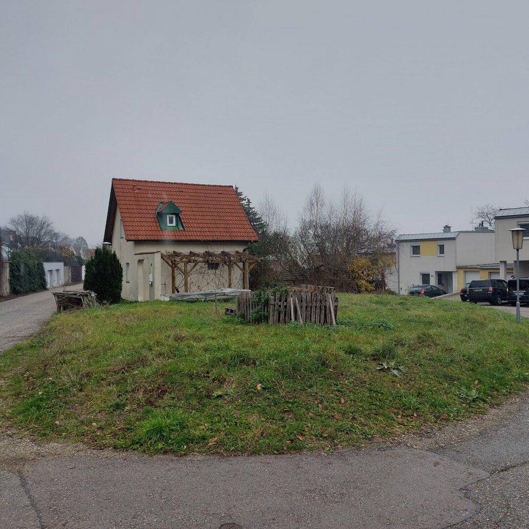 Unbebautes Grundstück mit einem kleinen Haus mit rotem Ziegeldach und einer Holzpergola im Hintergrund.