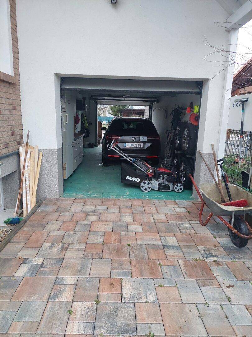 Einfahrt zur Garage mit gepflastertem Vorplatz, Auto und Gartengeräten.