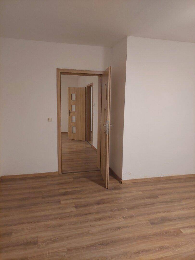 Leerer Raum mit Laminatboden und offener Holztür, die den Blick in einen weiteren Raum freigibt.