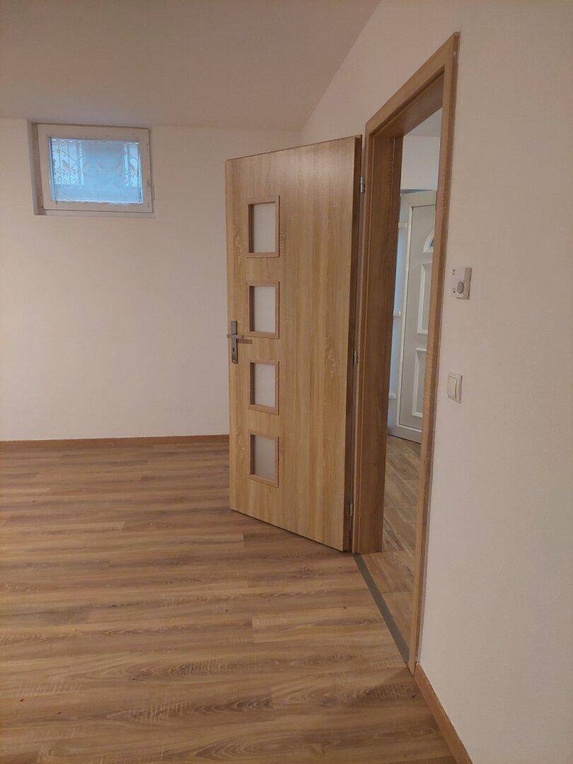 Leerer Raum mit Laminatboden und Fenster, eine Holztür führt in einen weiteren Bereich.