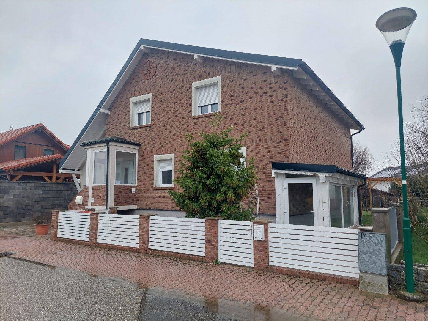Wohnhaus mit roter Ziegelfassade, weißem Zaun und einem kleinen Baum im Vorgarten.