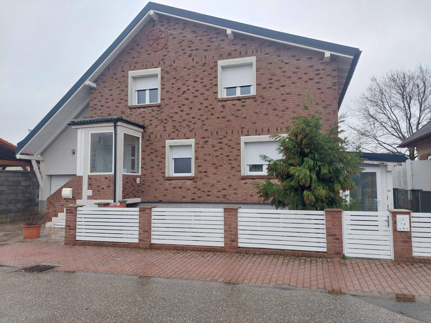 Vorderansicht des Ziegelhauses mit weißem Zaun, Erker und Rollläden an den Fenstern.