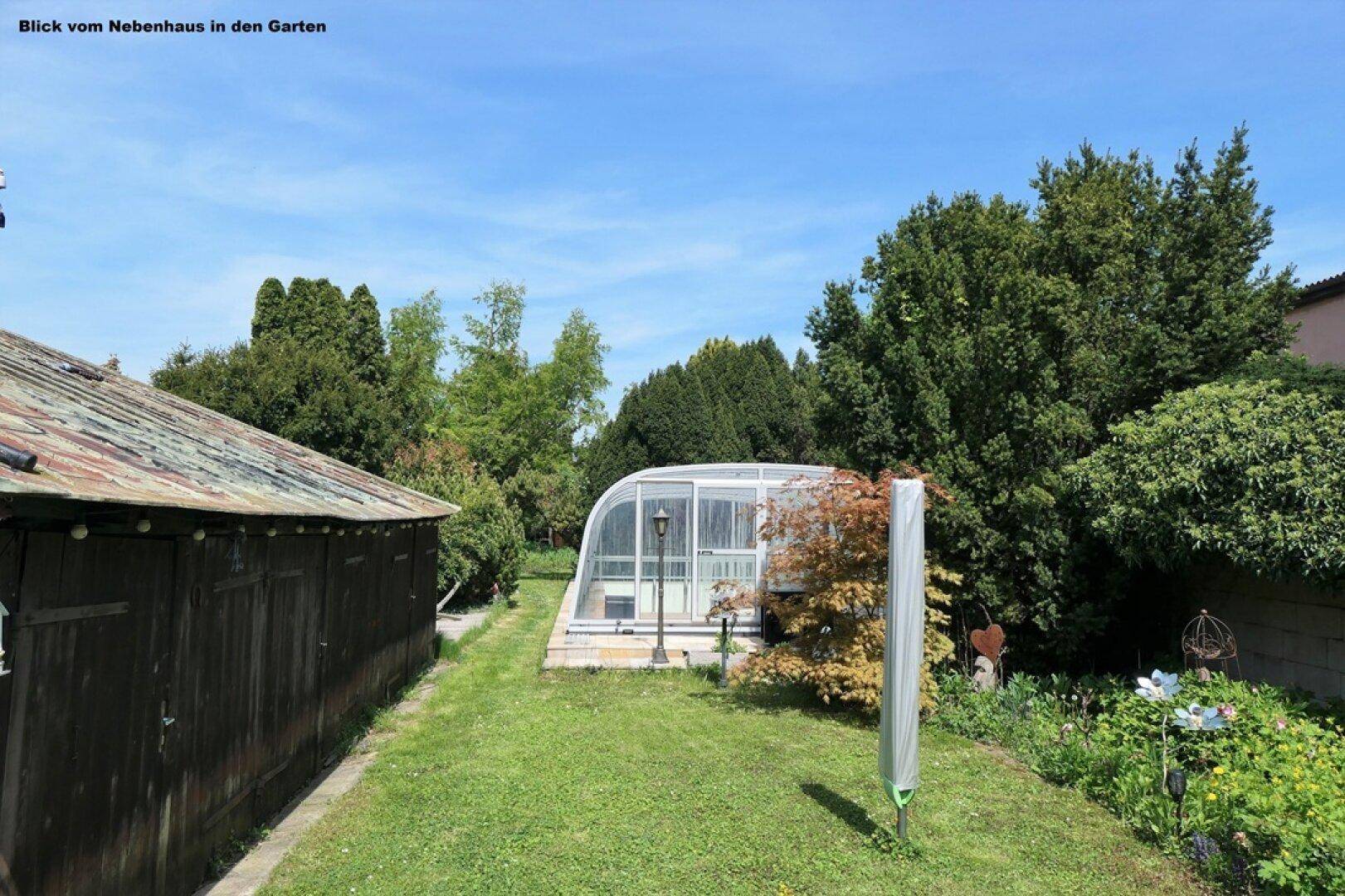 Weitläufiger Garten mit üppiger Vegetation, einem Schuppen und dem überdachten Pool im Hintergrund.