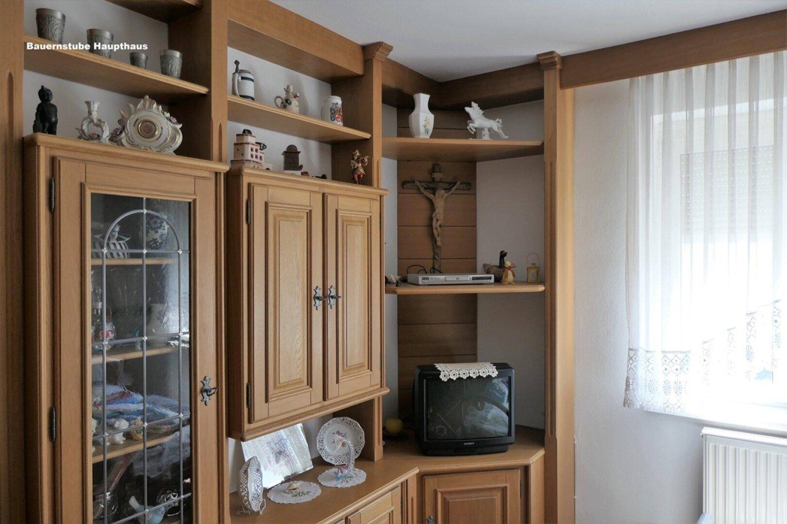 Traditionelle Bauernstube mit rustikalen Holzmöbeln, Regalen und einem kleinen Fernseher neben einem Fenster.
