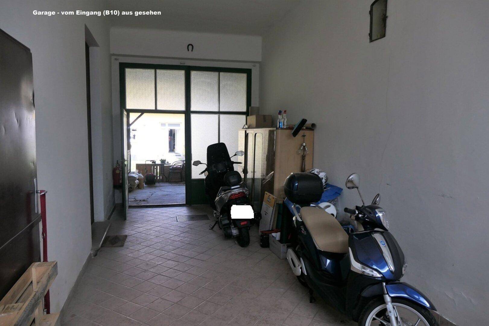Geräumige Garage mit Fliesenboden, zwei Rollern und Blick auf den Außenbereich durch eine Glastür.