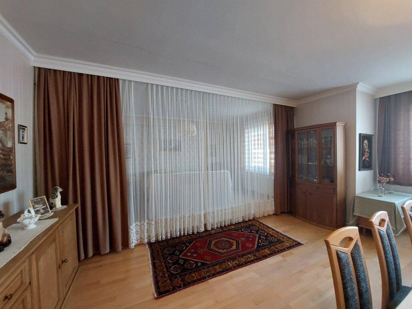 Wohnzimmer mit hellen Vorhängen, Holzmöbeln und einem gemusterten Teppich.