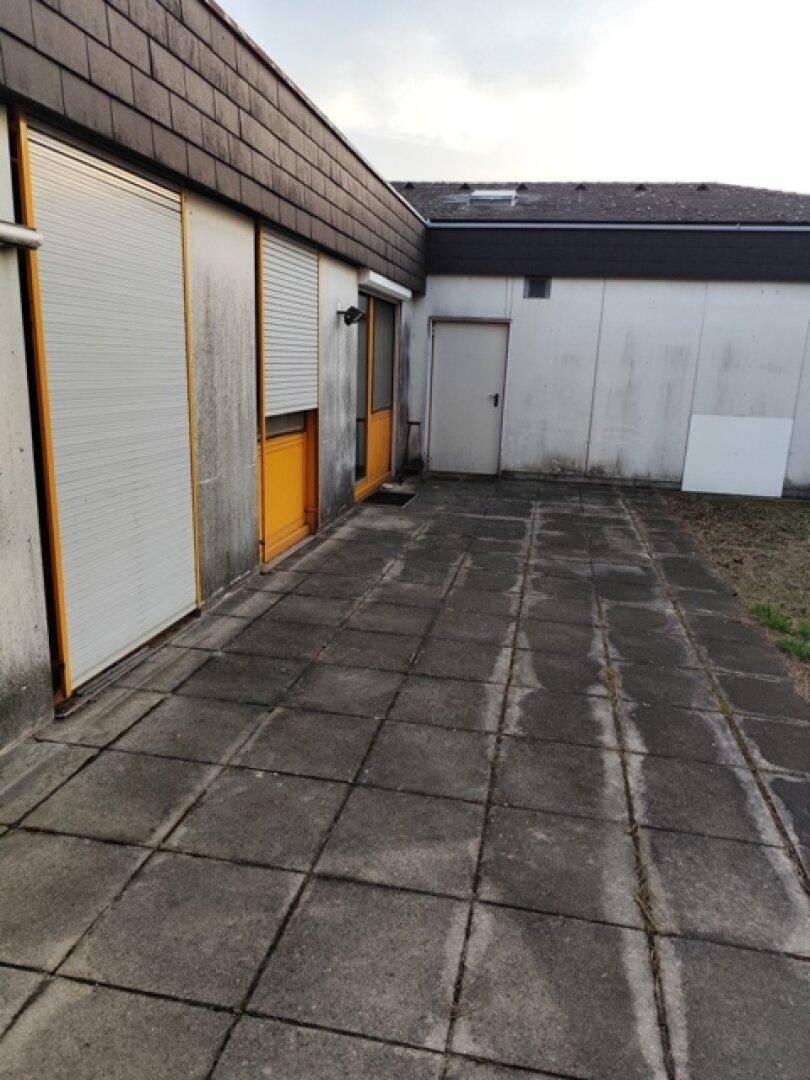 Gepflasterte Terrasse neben dem Gebäude mit weißen Wänden und Fenstern mit Rollläden.