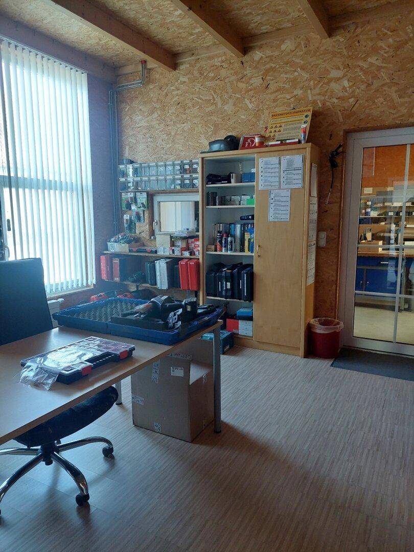 Funktionales Büro oder Werkstatt mit Schreibtisch, Regalen und Holzverkleidung, gut beleuchtet durch ein großes Fenster.