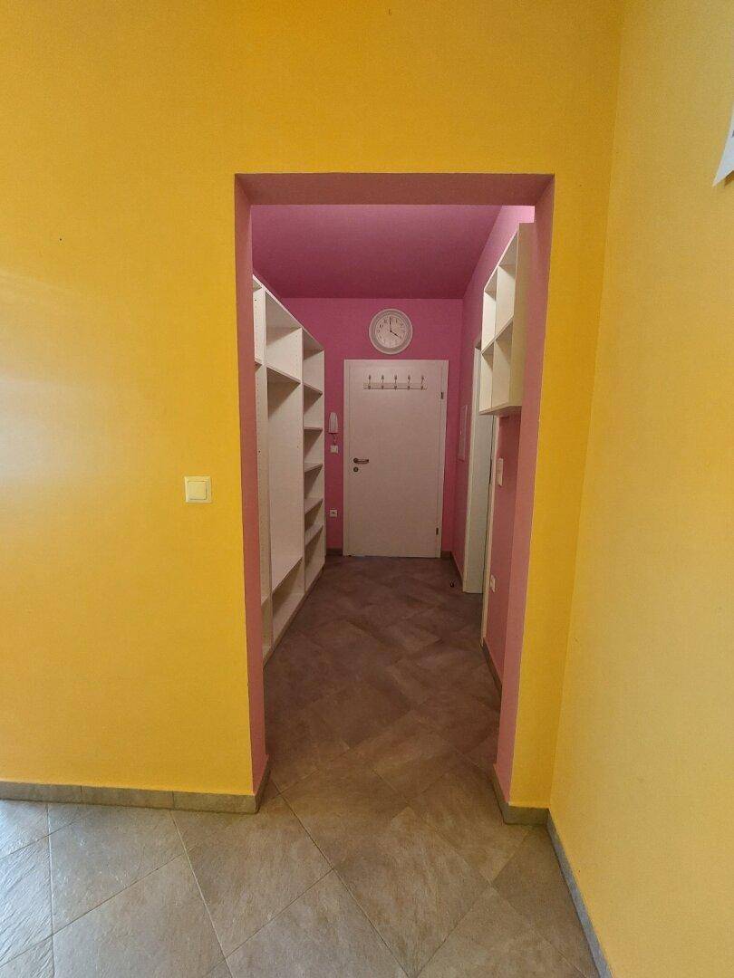 Blick aus einem gelben Raum in einen langen Flur mit rosa Wänden und weißen Regalen.