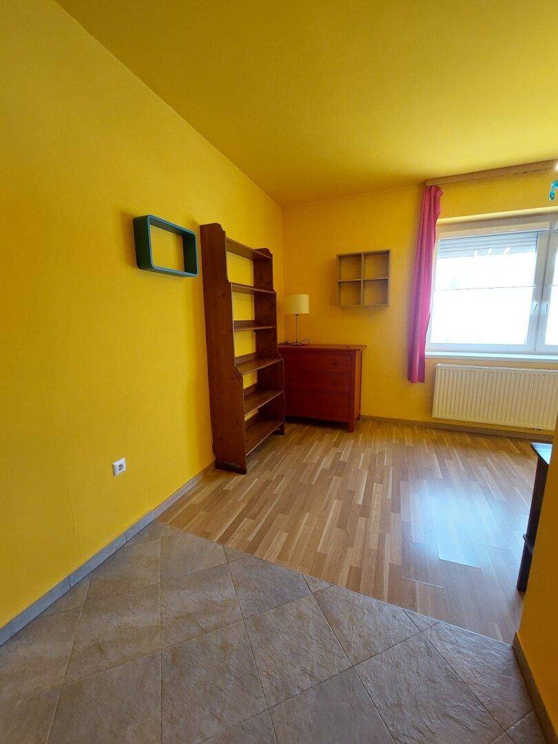 Helles Zimmer mit gelben Wänden, Holzfußboden und einem Fenster mit Heizkörper darunter.