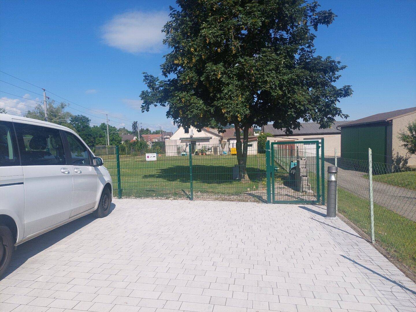 Außenparkplatz mit Pflastersteinen, umzäuntem Bereich und einem großen Baum.
