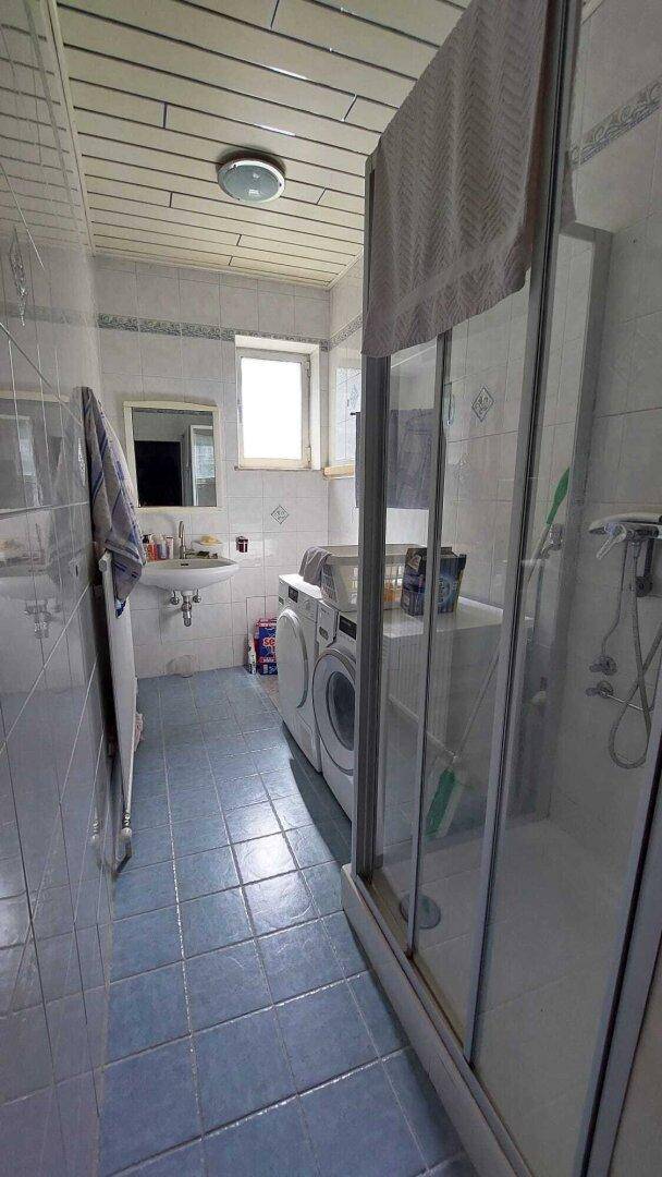 Badezimmer mit blauen Fliesen, Dusche und Waschmaschine, gut beleuchtet durch ein Fenster.