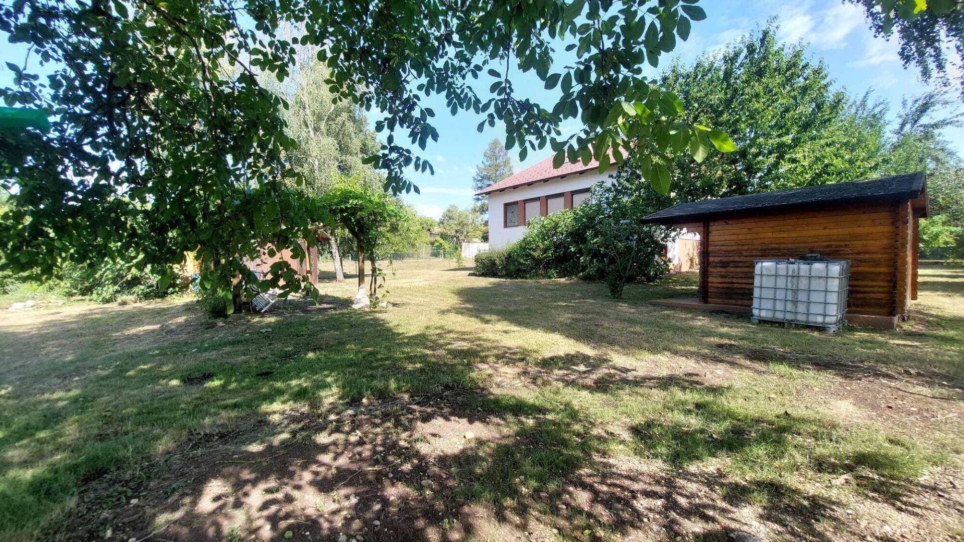 Gartenbereich mit einem Holzschuppen und dem Wohnhaus im Hintergrund unter Bäumen.