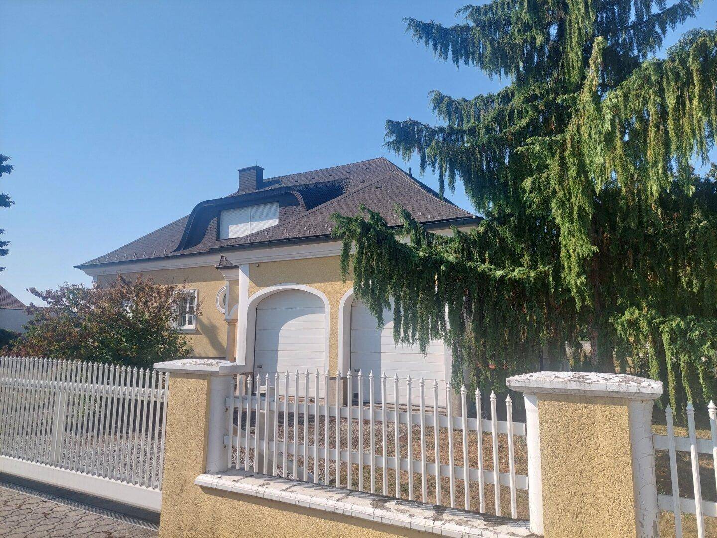 Attraktive gelbe Hausfassade mit zwei Garagentoren, umgeben von einem weißen Zaun und hohen Bäumen.