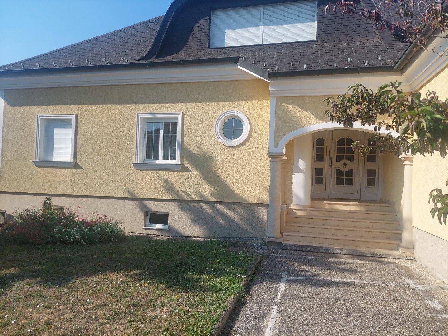 Eingangsbereich der Villa mit Säulen und Mansarddach unter strahlend blauem Himmel.