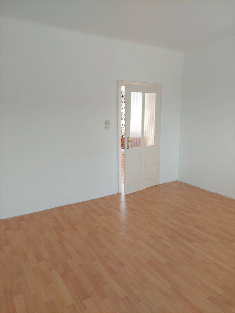 Leeres Zimmer mit Laminatboden und einer Tür, die in einen anderen Bereich führt.