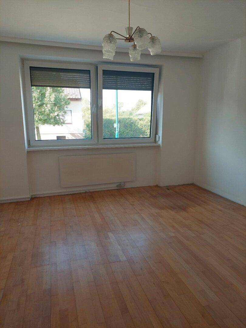 Geräumiges Wohnzimmer mit Parkettboden, großem Fenster und klassischer Deckenleuchte.