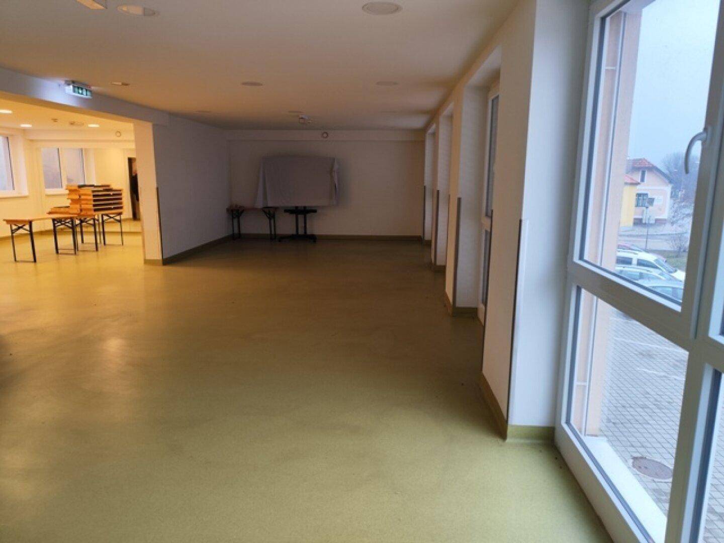 Heller Mehrzweckraum mit großen Fenstern, Blick nach draußen und funktionaler Einrichtung, bereit für Nutzung.