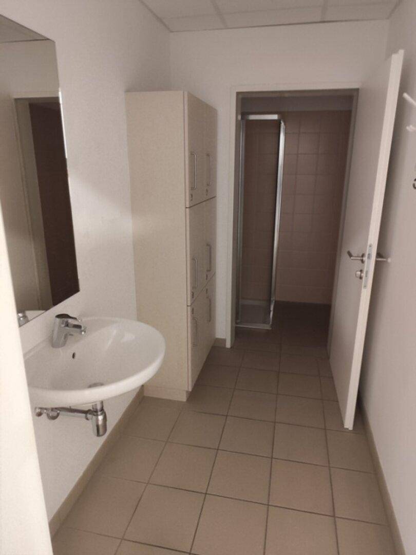 Umkleideraum mit Waschbecken, Spinden und Zugang zu einer Duschkabine, funktional und sauber.