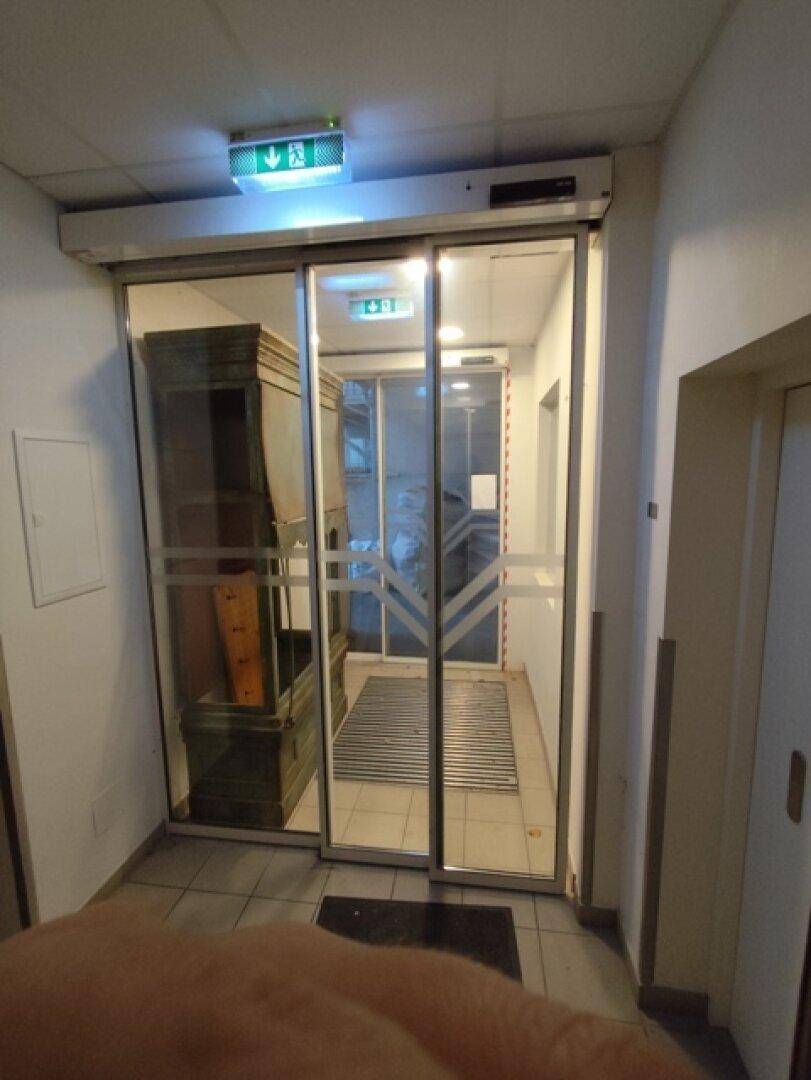 Eingangsbereich mit automatischen Glastüren und Fußmatte, führt in einen weiteren Flur.