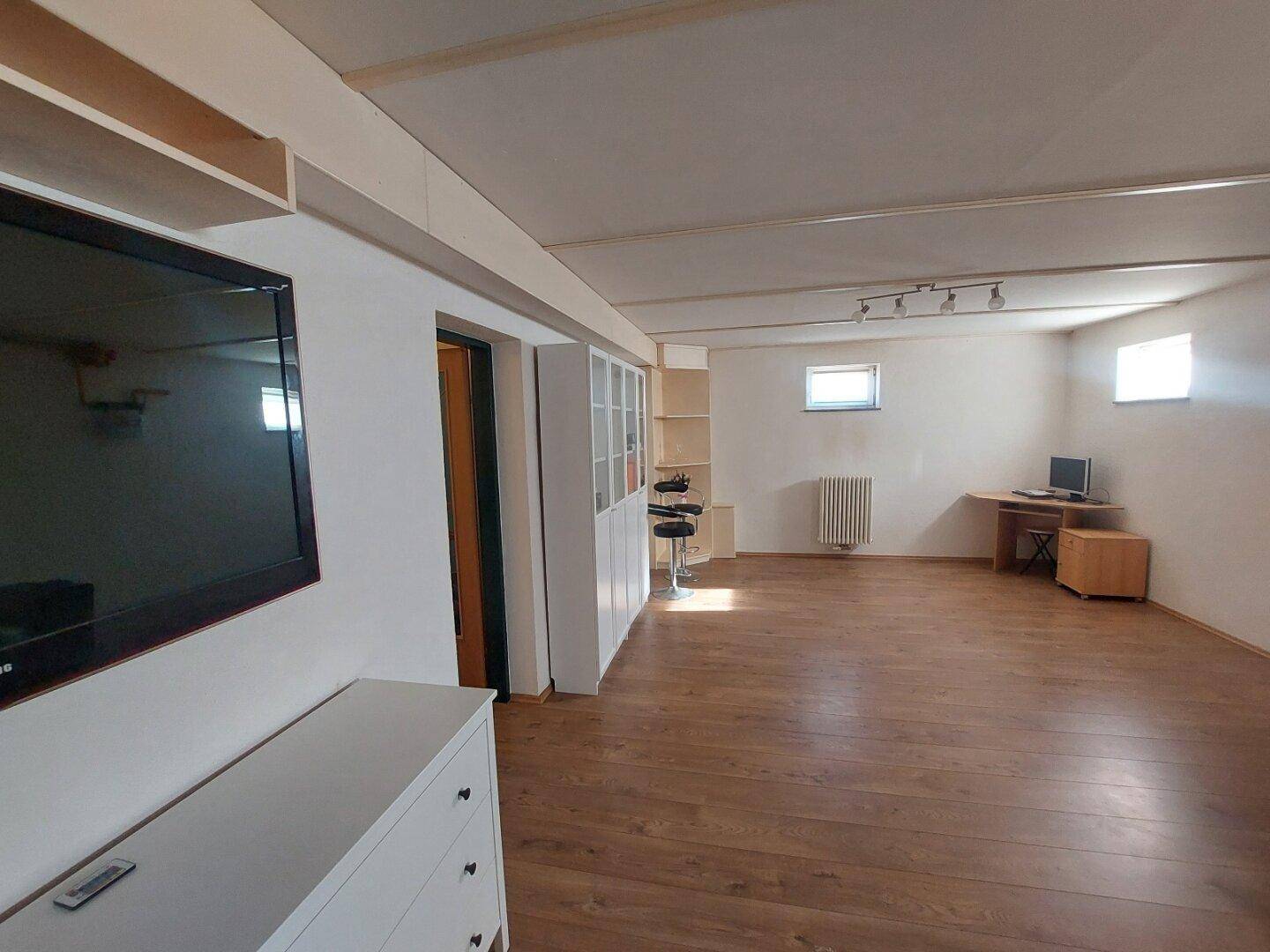 Großer Wohnraum im Untergeschoss mit Holzboden, Fenstern und TV-Bereich.
