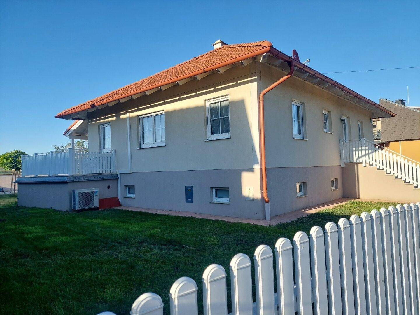 Seitenansicht des Hauses mit weißem Zaun, grünem Rasen und einer kleinen Terrasse im Hintergrund.