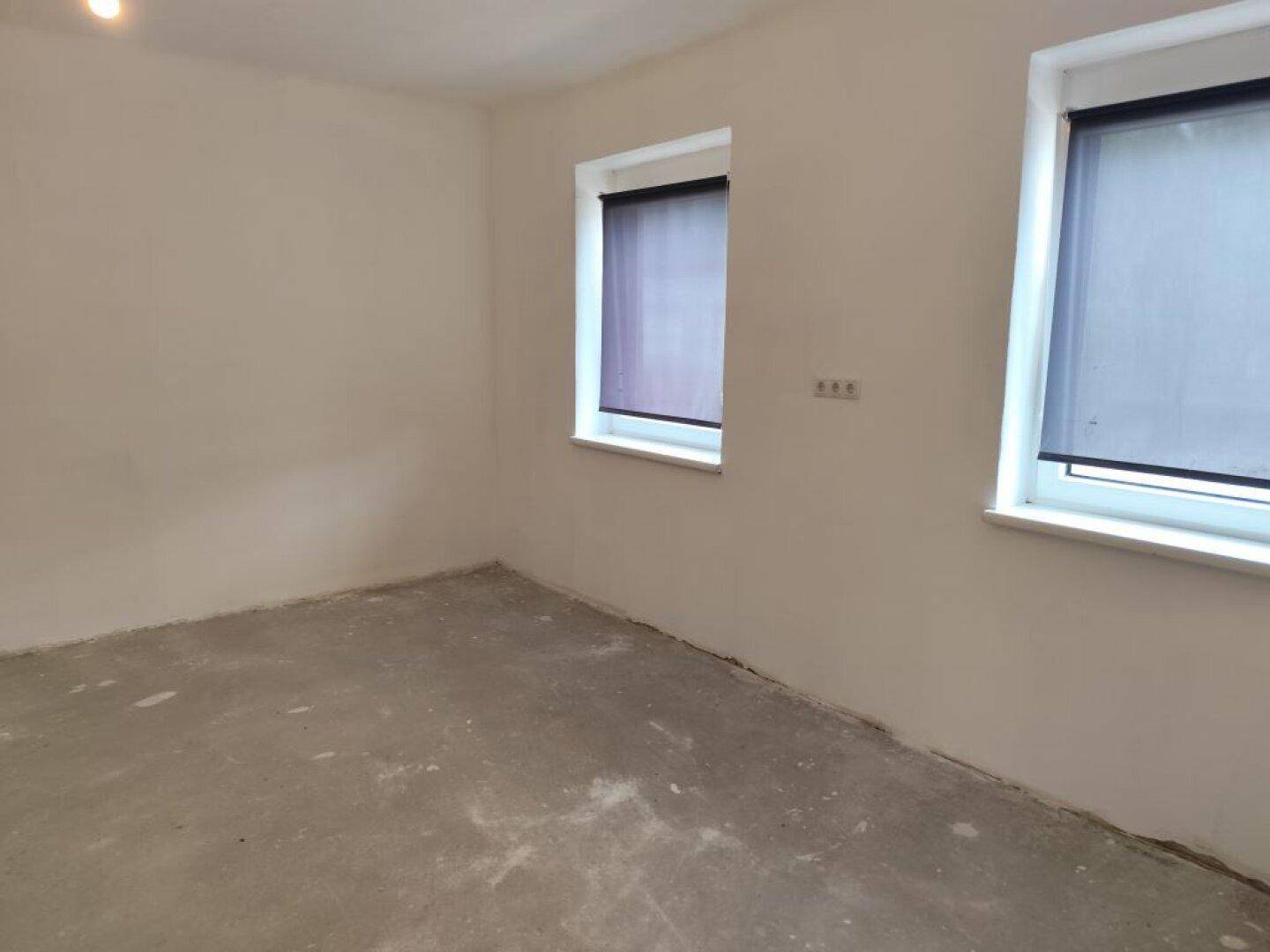 Heller Raum im Rohbau mit Betonboden, weißen Wänden und zwei Fenstern mit Rollos.