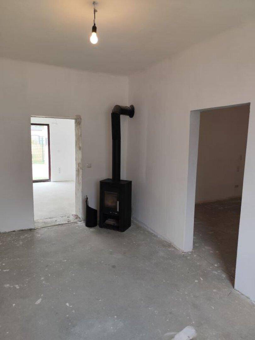 Wohnbereich im Rohbau mit Betonboden, weißen Wänden und einem modernen Holzofen.