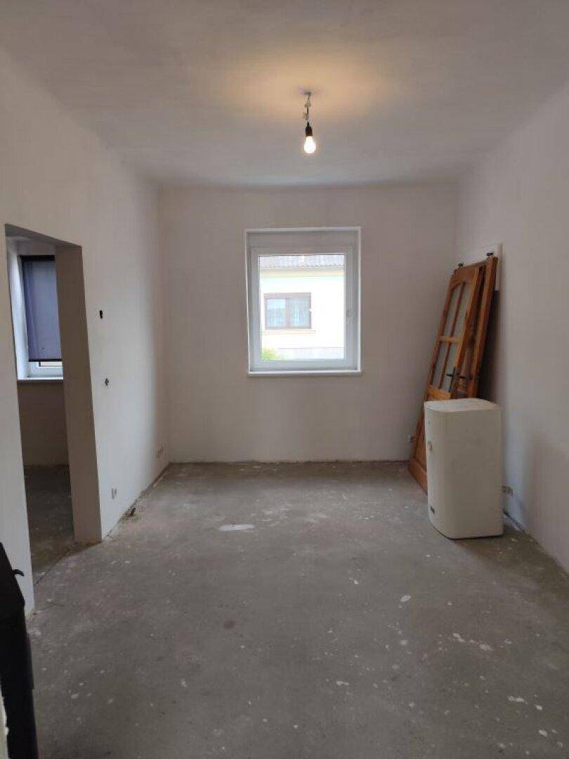 Heller, unfertiger Raum mit Betonboden, weißen Wänden und einem Fenster.