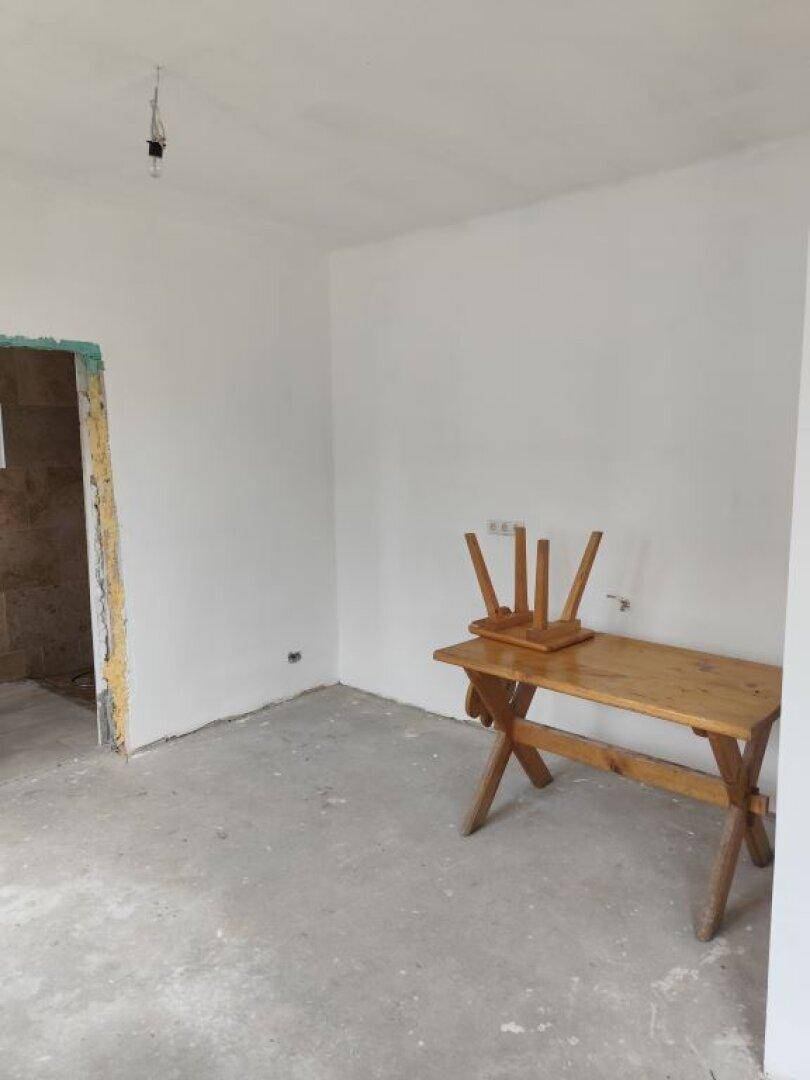 Unfertiger Raum mit einfacher Holzbank und Zugang zu einem gefliesten Bereich.