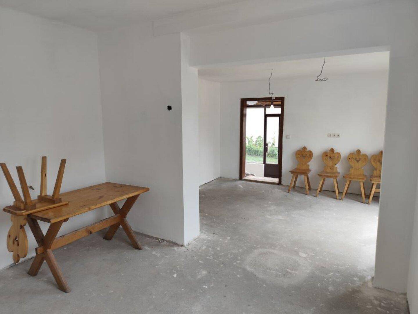 Offener Wohnbereich im Rohbau mit Betonboden, weißen Wänden und traditionellen Holzstühlen.