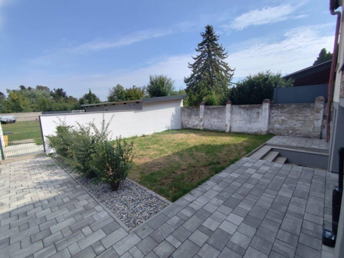 Gartenbereich mit Rasen und gepflastertem Weg, umgeben von einer Mauer und Bäumen.