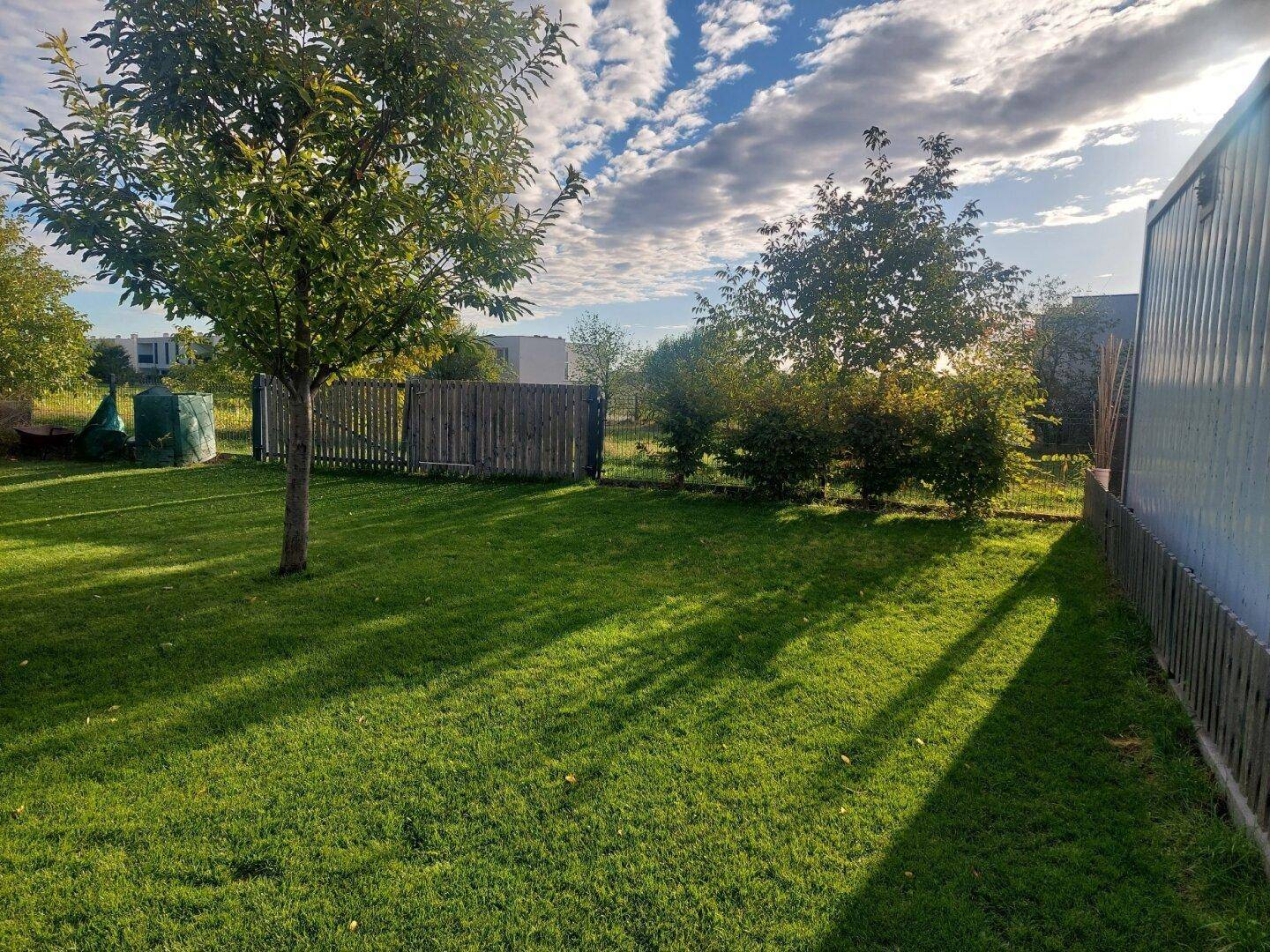 Gepflegter Garten mit Rasenfläche, Holzzaun und Bäumen unter blauem Himmel.